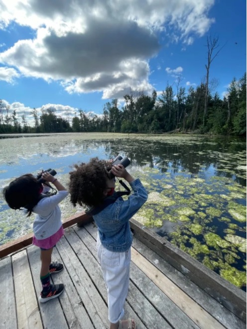 Two kids next to a lake look up at the sky with binoculars.